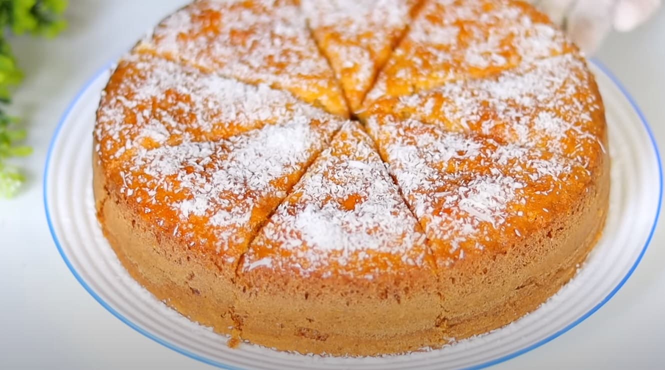 Um bolo dourado e fofinho repousa sobre uma bandeja rústica, salpicado com coco ralado fresco, com uma fatia recém-cortada revelando sua textura macia e úmida. Ao fundo, ingredientes como coco, farinha e maizena estão elegantemente dispostos,